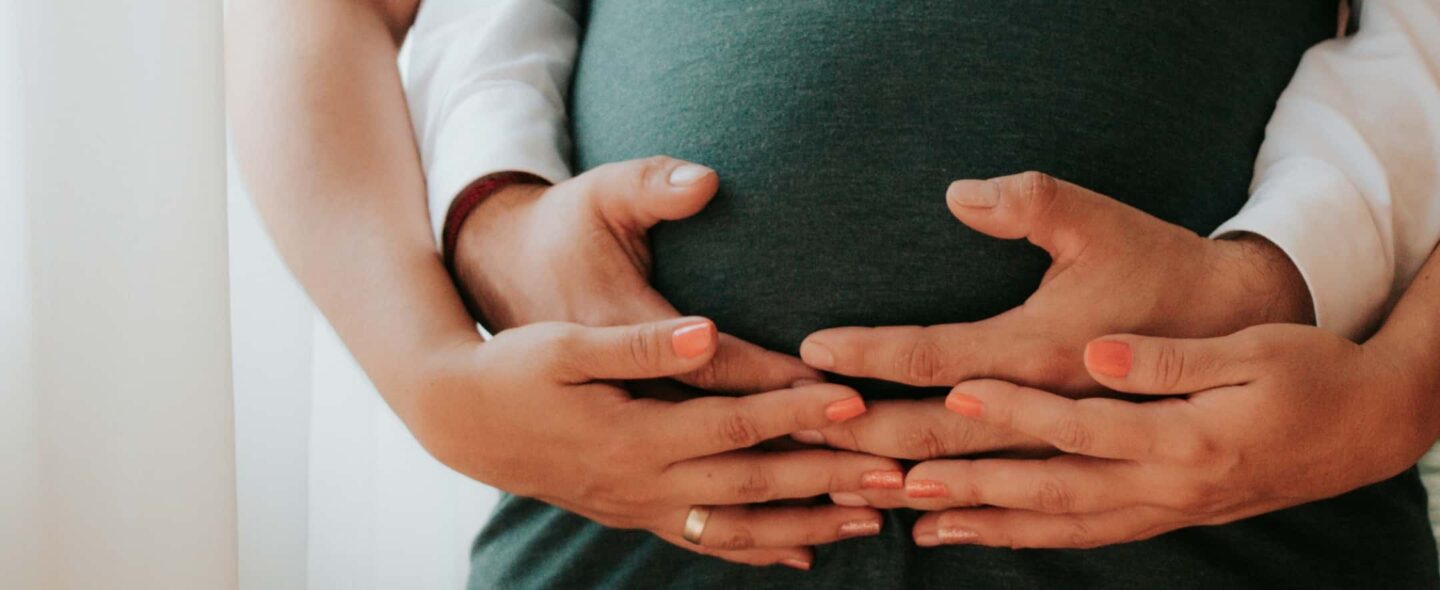 Händer på en gravid mage, två par händer håller runt magen.